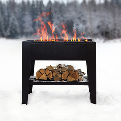 A large outdoor grill for barbequing food. It has fire blazing, with a pile of wood underneath. Snow has settled on the ground and surrounding area. 